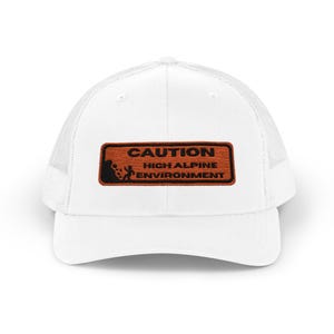 May include: White trucker hat with a mesh back and a rectangular patch. The patch is brown with the words "CAUTION HIGH ALPINE ENVIRONMENT" in black. A small graphic of a person and falling rocks is on the left side of the patch.