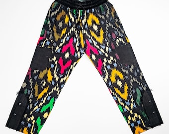 May include: Black patterned pants with a colorful geometric design in yellow, pink, green, and blue. The pants have black pockets and a black drawstring waistband. The cuffs have black button details.