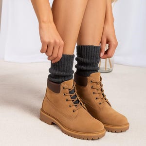 Könnte beinhalten: Hellbraune Schnürstiefeletten mit passenden Schnürsenkeln, einem dunkelbraunen gepolsterten Schaftrand und einer dicken, profilierten Gummisohle. Die Stiefel werden mit dunkelgrauen, gerippten Strickstrümpfen getragen, die über den Schaft hinausragen.