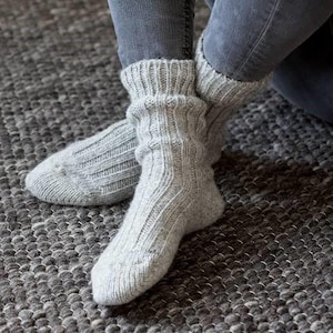 Könnte beinhalten: Ein Paar hellgraue Stricksocken mit gerippten Bündchen und einem strukturierten, vertikalen Muster. Die Socken werden über grauen Jeans getragen und liegen auf einem grau strukturierten Teppich, was Gemütlichkeit und Wärme suggeriert.