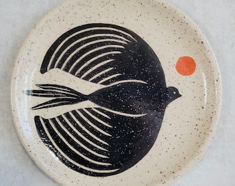 May include: A round ceramic plate with a speckled cream-colored glaze. The center features a large, stylized black bird in flight with detailed wing feathers. A small, solid orange circle is positioned to the right of the bird's head.