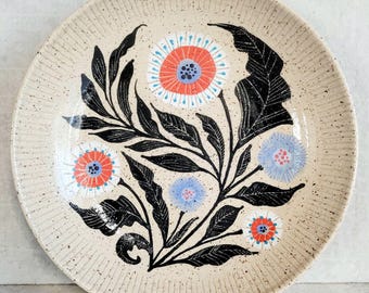May include: A round ceramic plate with a speckled cream-colored base and a ribbed rim. The center features a hand-painted botanical design with black leaves and five stylized flowers in shades of coral, light blue, and white with dark centers.