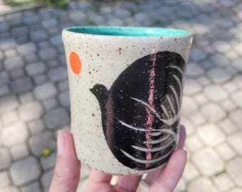 May include: A cylindrical ceramic cup with a speckled off-white exterior and a bright turquoise interior rim. The side features a large black bird silhouette with white etched feather details and a small solid orange circle representing the sun.
