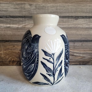 May include: A cream-colored ceramic vase with a rounded body and a narrow neck. The surface features dark blue illustrations of two birds facing each other, separated by a central white flower and a small white circle near the top rim.