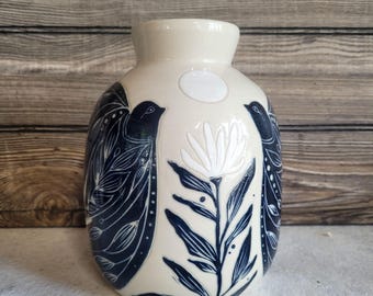 May include: A cream-colored ceramic vase with a rounded body and a narrow neck. The surface features dark blue illustrations of two birds facing each other, separated by a central white flower and a small white circle near the top rim.