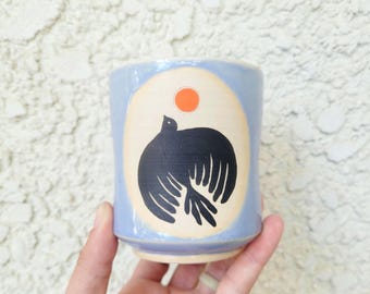 May include: A light blue ceramic cup featuring a central cream-colored oval panel. Inside the oval, a black bird is depicted in flight beneath a small, solid orange circle representing the sun. The cup has a smooth, glossy finish and a cylindrical shape.