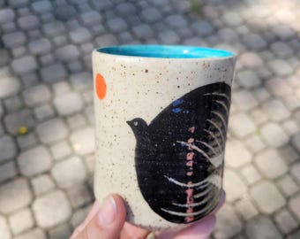 May include: A cylindrical ceramic cup with a speckled cream-colored exterior featuring a large black bird illustration, a small orange circle, and a bright turquoise interior rim. The cup has a smooth, straight-sided design without a handle.