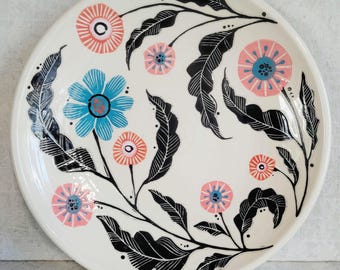 May include: A round ceramic plate with a cream-colored base featuring a hand-painted botanical design. The pattern includes black outlined leaves and various flowers in shades of bright blue, soft pink, and coral orange with dotted centers.