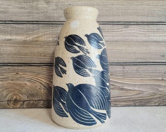 May include: A ceramic vase with a speckled cream-colored base and a repeating pattern of dark blue birds in flight. The vase features a tapered cylindrical shape with a narrow neck and a wide, rounded opening at the top, showcasing a smooth finish.