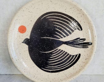 May include: A round ceramic plate with a speckled cream-colored glaze. The center features a large, stylized black bird in flight with detailed wing feathers. A small, solid orange circle is positioned to the left of the bird's head.