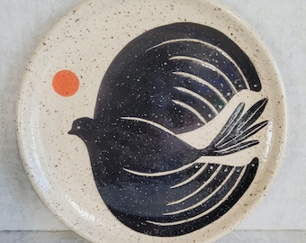 May include: A round ceramic plate with a cream-colored base featuring dark brown speckles. The center displays a large, stylized black bird in flight with white line details on its wings and tail, accompanied by a small, solid orange circle.
