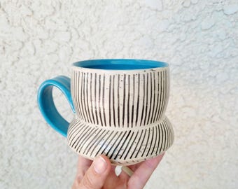 May include: A ceramic mug with a bright blue interior and handle. The exterior features a cream-colored base with a pattern of vertical black lines on the upper section and diagonal black lines on the wider, rounded lower base section.