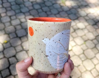 May include: A cylindrical ceramic cup with a speckled cream-colored glaze, featuring a painted white bird silhouette and a small solid orange circle. The interior rim of the cup is finished in a vibrant orange glaze, contrasting with the exterior.