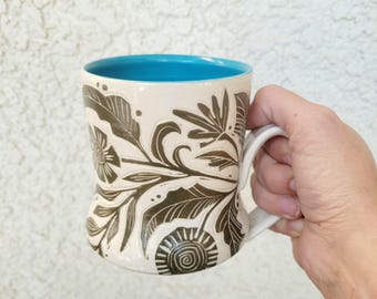 May include: A ceramic mug with a cream-colored exterior featuring a dark brown botanical relief pattern of leaves and flowers. The interior of the mug is a bright turquoise blue, and it includes a curved handle on the side for easy gripping.
