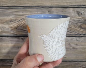 May include: A ceramic mug with a light beige exterior and a smooth, light blue interior. The outer surface features a white, etched graphic of a bird with detailed wing feathers and a small, solid orange circle positioned near the bird's head.