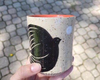 May include: A cylindrical ceramic cup with a speckled cream-colored exterior featuring a large black bird illustration and a small white circular accent. The interior of the cup is a solid, vibrant orange color, and the base has a ribbed texture.