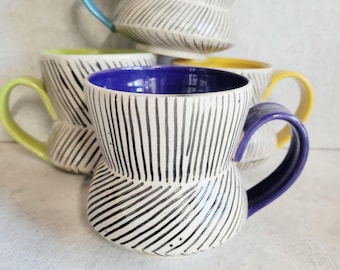 May include: A collection of ceramic mugs featuring a white exterior with black vertical and diagonal etched lines. Each mug has a distinctively colored handle and interior, including shades of bright blue, lime green, yellow, and deep purple.