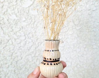 May include: A small, bulbous ceramic vase with a cream-colored glaze and vertical ribbed texture. The vase features hand-painted black dots and horizontal lines around its midsection and is filled with a cluster of dried, light beige baby's breath.