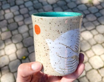 May include: A speckled ceramic tumbler with a light beige exterior and a vibrant turquoise interior. The exterior features a small orange circle and a large white abstract shape with etched line details, creating a textured, hand-crafted appearance.