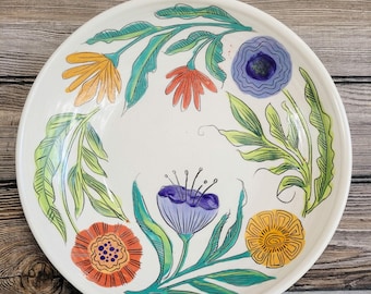 May include: A round white ceramic plate featuring a hand-painted botanical design. The illustration includes various stylized flowers in shades of orange, yellow, and purple, connected by long, flowing green leaves and stems across the surface.