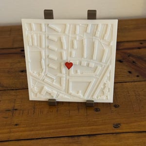 Può includere: Una mappa bianca quadrata stampata in 3D con un cuore rosso che segna una posizione. La mappa è supportata da due supporti marroni. La mappa mostra una planimetria dettagliata, perfetta per un regalo personalizzato o l'arredamento della casa.