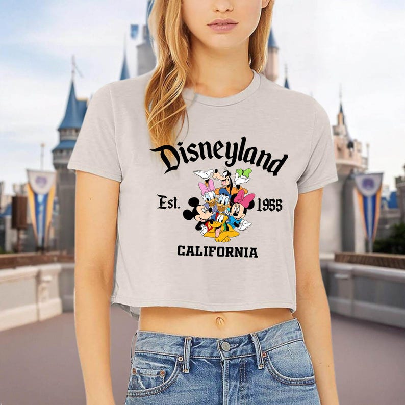 May include: A light gray crop top featuring the Disneyland logo with Mickey Mouse, Donald Duck, Goofy, and other Disney characters. The text reads "Disneyland Est. 1955 California" in black lettering. The shirt is paired with blue jeans.