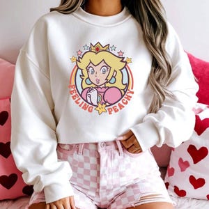 May include: White sweatshirt featuring a cartoon illustration of Princess Peach with the text "Feeling Peachy". The design includes a rainbow, stars, and a crown. The sweatshirt is paired with pink checkered shorts.