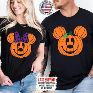 May include: Two black t-shirts featuring orange pumpkin graphics shaped like mouse heads. The left shirt displays a pumpkin with a purple polka-dot bow, while the right shirt shows a pumpkin with a green leaf. Both have smiling faces.