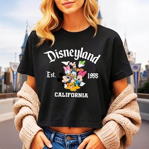 May include: Black t-shirt featuring the Disneyland logo with Mickey Mouse and friends. The shirt includes the text "Est. 1955 CALIFORNIA" in white. The design showcases classic Disney characters in a colorful graphic.