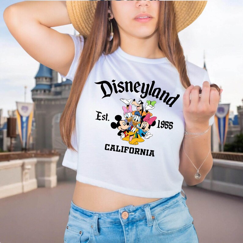 May include: White crop top featuring the word "Disneyland" in a script font, with the text "Est. 1955 CALIFORNIA" and cartoon characters. The shirt is paired with blue jeans.