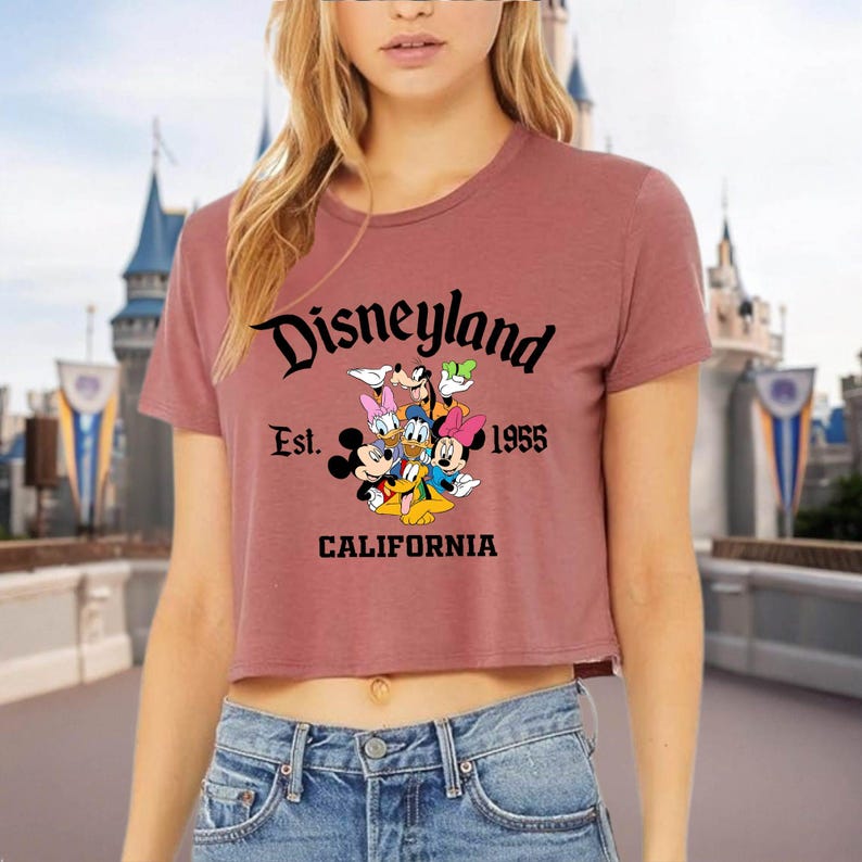 May include: A dusty rose crop top featuring the Disneyland logo with Mickey Mouse, Donald Duck, Goofy, Minnie Mouse, and Daisy Duck. The text reads "Disneyland Est. 1955 California". The shirt is paired with blue jeans.