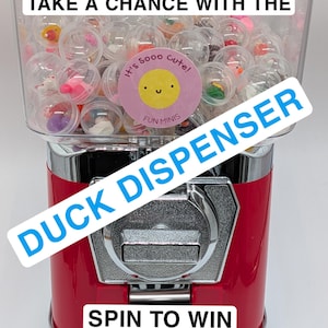 May include: A red and chrome duck dispenser with the text "TAKE A CHANCE WITH THE DUCK DISPENSER SPIN TO WIN." The dispenser contains clear capsules with miniature duck figurines. A pink sticker reads "It's sooo cute! FUN MINIS."