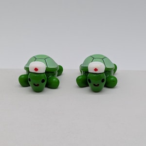May include: Two small green turtle figurines, each wearing a white nurse cap with a small red cross symbol on the front. The turtles have smooth, glossy green bodies, dark eyes, and a smiling mouth, with a textured light green shell pattern.