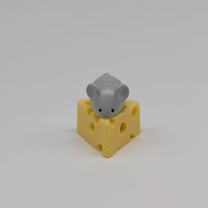 May include: A small, gray mouse figurine with black eyes sits atop a yellow, Swiss cheese-shaped object. The cheese has several round holes. The background is a plain, light gray.