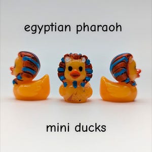 May include: Three miniature orange duck figurines, each adorned with a pharaoh headdress in blue and brown. The text "egyptian pharaoh" and "mini ducks" are displayed above and below the ducks.
