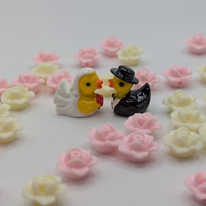 May include: A pair of miniature duck figurines dressed as a bride and groom, surrounded by small pink and cream-colored rose decorations. The bride duck wears a white veil, and the groom duck is in a black tuxedo and top hat. The figurines are yellow with orange beaks.