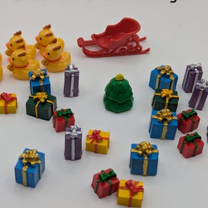May include: A collection of miniature decorative items, including yellow ducklings, a red sleigh, a green Christmas tree, and colorful gift boxes with gold bows. The text "decorate a mini village!" is at the top.