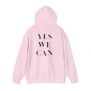 Pode incluir: Um moletom rosa claro com as palavras "YES WE CAN" impressas em preto nas costas. O moletom tem capuz e mangas compridas e é feito de um material macio.