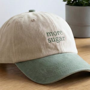 May include: A beige baseball cap with a green brim. The phrase "more sugar" is embroidered in green on the front of the cap. The cap is sitting on a wooden surface.