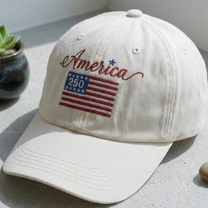 May include: Cream-colored baseball cap with the word "America" embroidered in red script, along with a small American flag patch. The flag patch includes the number "250". The cap has a curved brim and a button on top.