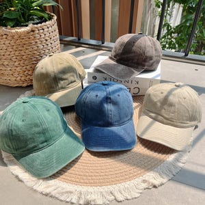 May include: Five baseball caps in various colors: green, blue, tan, and gray. The caps are made of a soft fabric and have a classic six-panel design with a curved brim. The caps are arranged on a round, fringed mat.