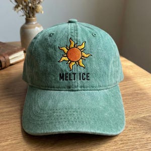 May include: A green baseball cap with an embroidered sun design in orange and yellow. The text "MELT ICE" is embroidered below the sun. The cap is made of a soft, worn material.