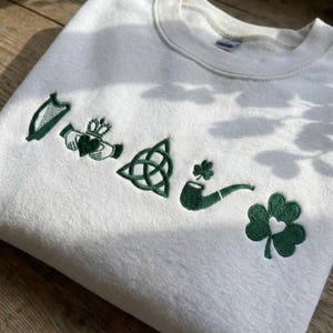 May include: White sweatshirt with green embroidered Irish symbols, including a harp, Claddagh ring, Celtic knot, smoking pipe, and shamrock. The crew neck sweatshirt is made of a soft material.