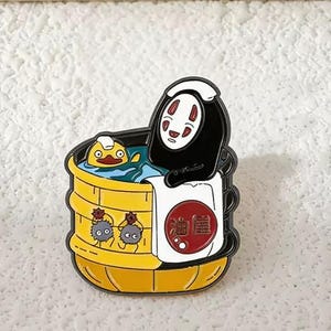 Può includere: Una spilla smaltata colorata con personaggi di un popolare film d'animazione. La spilla raffigura una figura nera con una maschera bianca in una vasca di legno gialla con un'anatra e due folletti della fuliggine. La vasca ha un asciugamano bianco con testo giapponese.