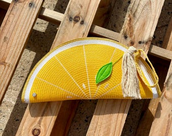 Puede incluir: Un bolso de mano semicircular con forma de rodaja de limón, que presenta una superficie texturizada de color amarillo brillante con líneas radiales blancas, una pequeña aplicación de hoja verde y una borla beige en una correa amarilla.