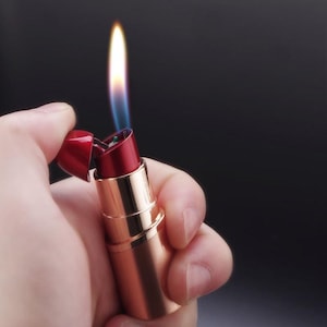 May include: A red lipstick-shaped lighter with a gold-colored base. The lighter is open, with a flame emitting from the top. The background is a dark, solid color.