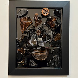 May include: A framed display featuring a collection of brown glass perfume bottles and decorative elements. The frame is black, and the arrangement includes a Versace Eros bottle, a copper-colored coin, and various geometric shapes.