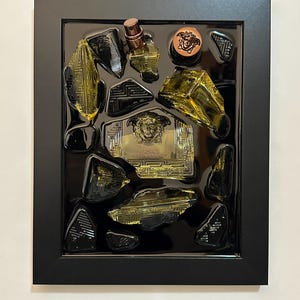 May include: A framed artwork with gold and black decorative elements. It features a perfume bottle, a round emblem, and geometric shapes, set against a black background. The frame is black. The artwork is presented in a black frame.