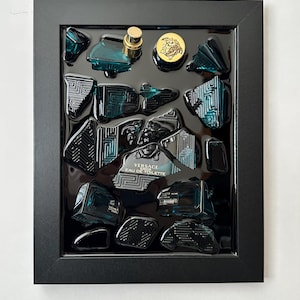 May include: A framed artwork with fragments of a teal perfume bottle, a gold cap, and a gold emblem. The fragments are set on a black background within a black frame. The text "VERSACE EAU DE TOILETTE" is visible on the bottle.