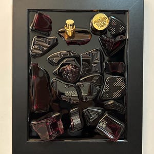 May include: A framed artwork featuring fragments of a dark red perfume bottle and cap, arranged on a black background. The gold cap and a gold emblem are visible. The text "VERSACE EROS FLAME EAU DE PARFUM" is also visible.